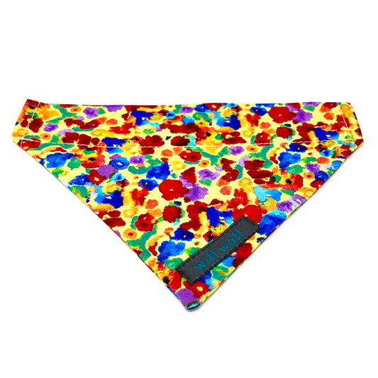 A dog collar bandana in 100% cotton fabric made up of a light yellow background adorned with irregular, abstract blotches in red, blue, green, purple, and occasional orange. 