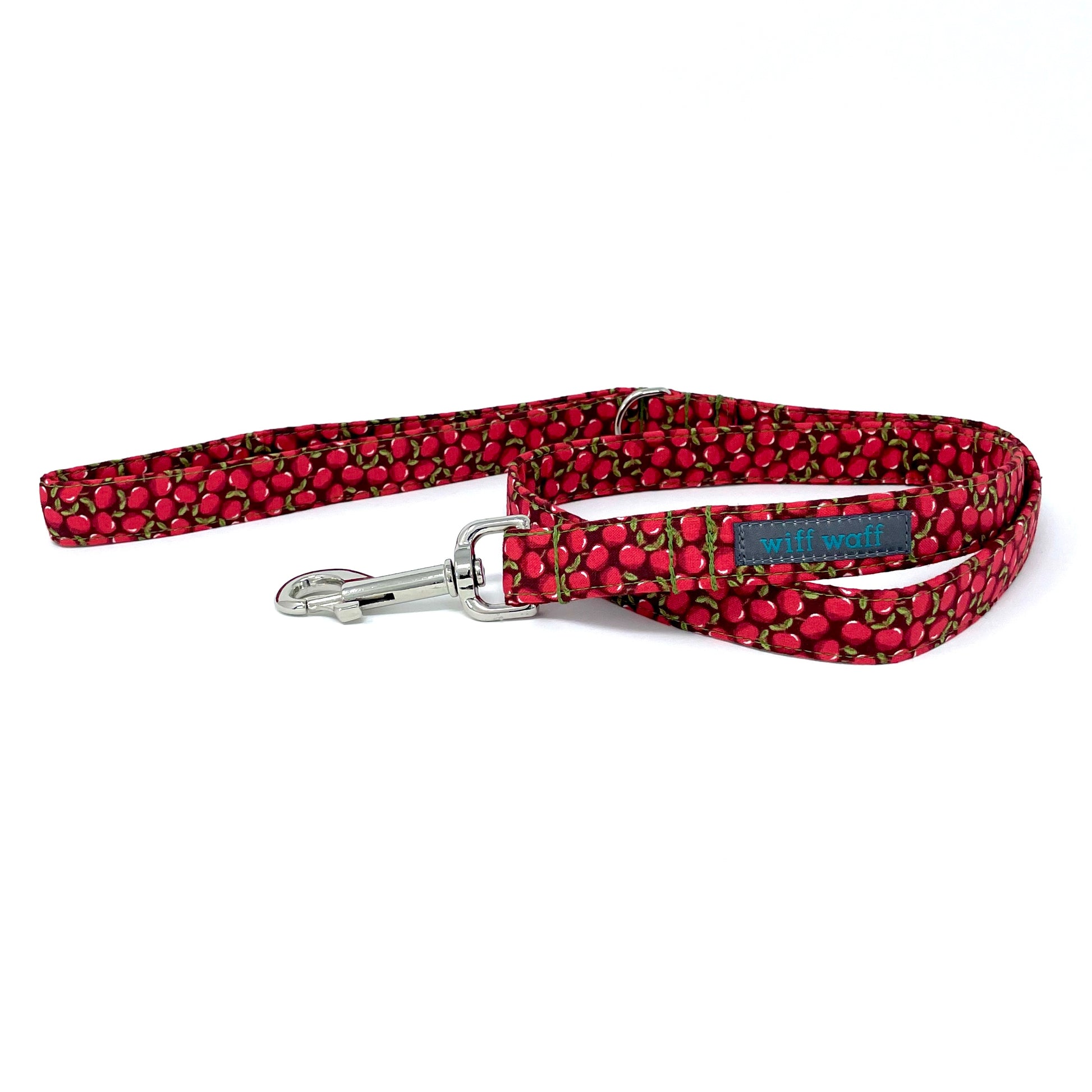 A coiled up handcrafted fabric dog lead in a red apple print on a dark background with green stitching and chrome clip and d ring. Handcrafted by wiff waff designs in nottingham
